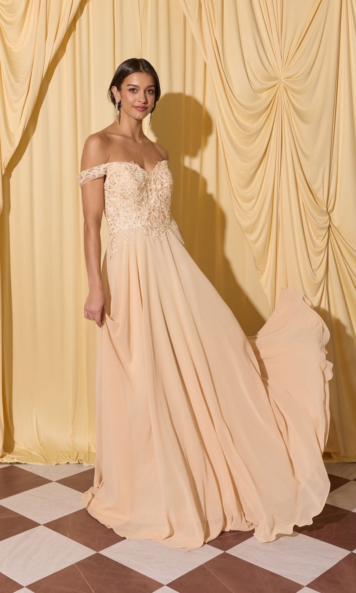 A woman in the Long Prom Dress: Eureka EK211 in Champagne stands on a checkered floor, lifting her flowing skirt before beige curtains.